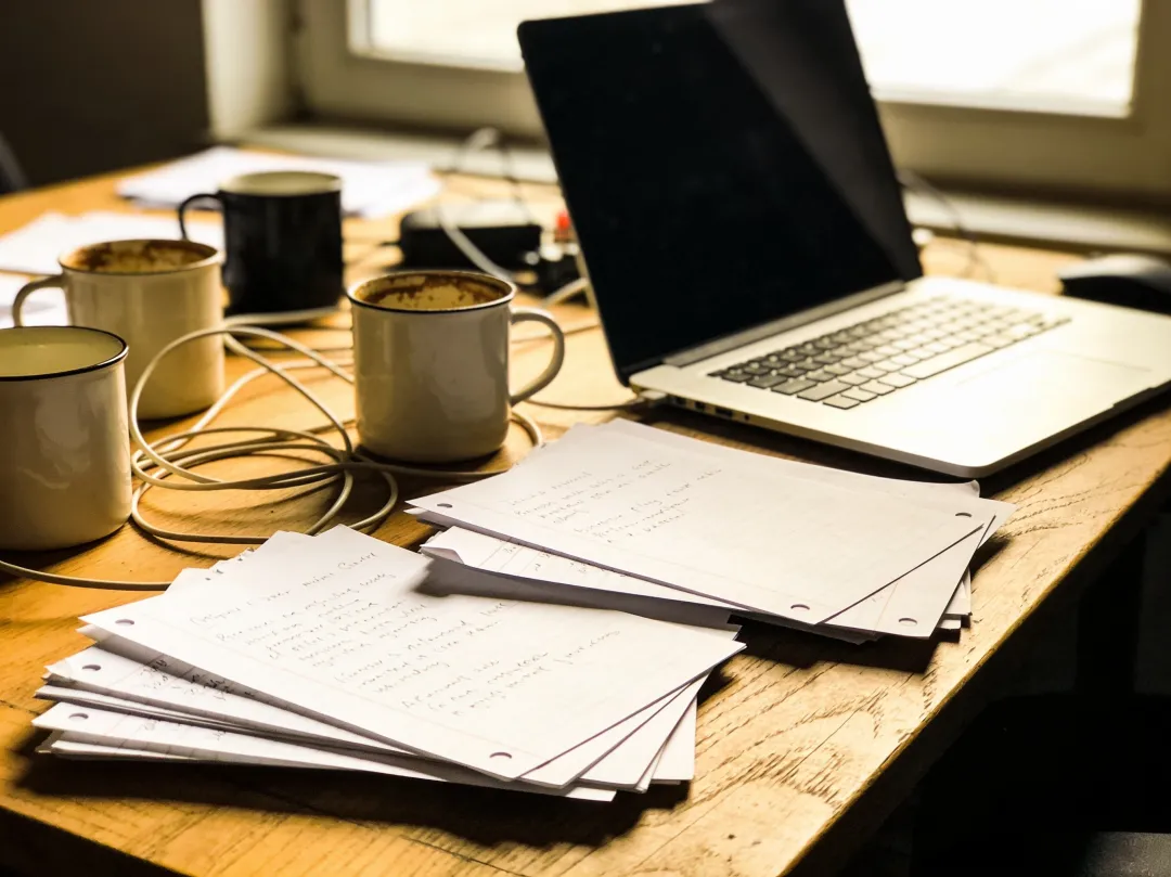 Original photo — a desk cluttered with handwritten papers, mugs, and cables around a laptop, before the AI edit