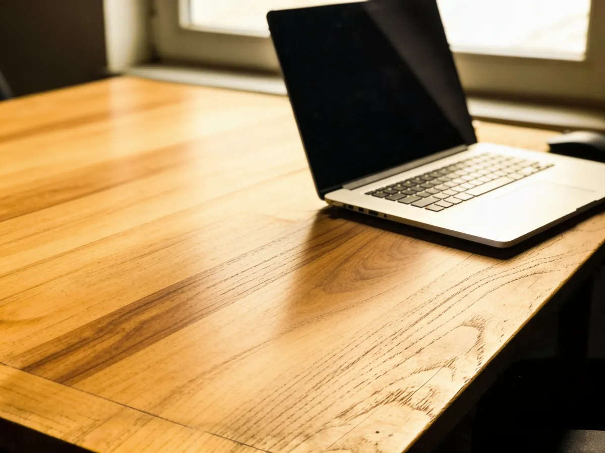 AI photo edit — cluttered desk cleared to a single laptop on a walnut wood surface by a window